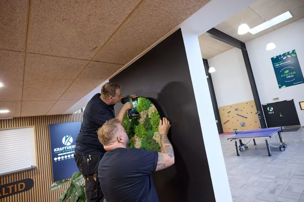 To mænd monterer en rund mosvægdekoration på en sort væg i Kraftværket Videbæk