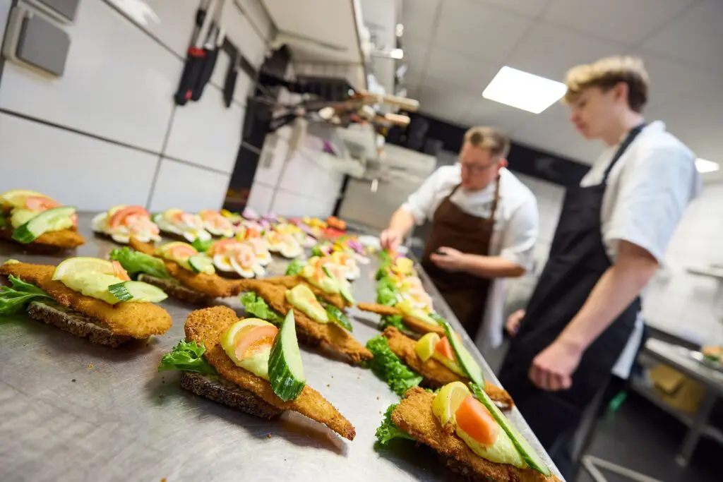 Kokke anretter frisksmurt smørrebrød med fiskefilet og grønt på et stålbord i et industrikøkken