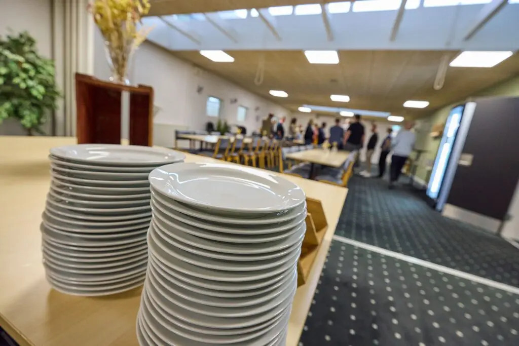 Stabler af hvide tallerkener på et bord i forgrunden med gæster i baggrunden i møde- og selskabslokale.