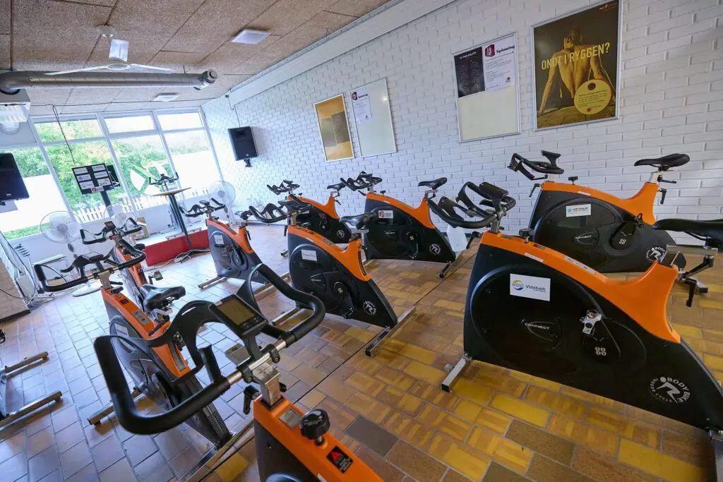 Rækker af orange og sorte spinningcykler i et lyst træningslokale med klinkegulv