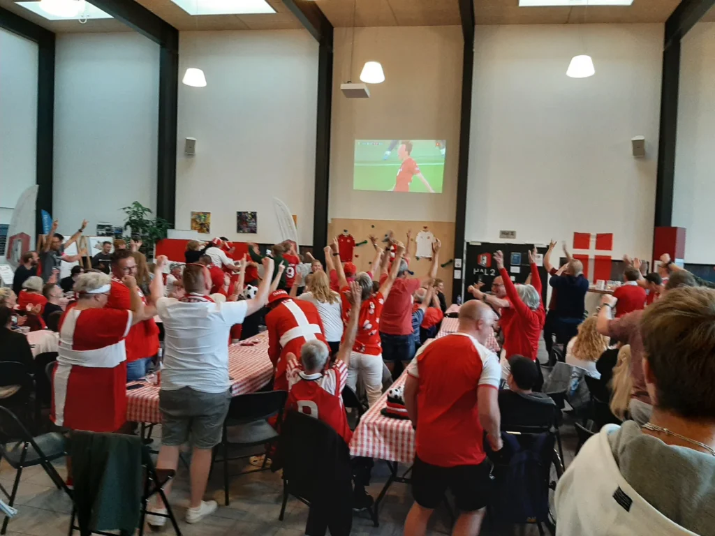 Glade tilskuere i røde og hvide trøjer jubler foran storskærm til sportsarrangement i Kraftværket Videbæk.